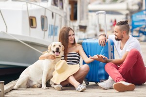 A young married couple,a man with a beard and a pregnant woman,brunette,long straight hair, with a friend,a Labrador dog,awaiting departure on a journey,sitting on the dock with a big blue suitcase waiting for the ship